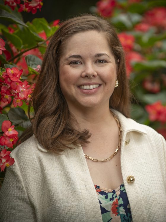 A person in a white jacket smiling in front of red flowering bushes.