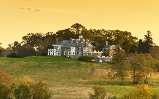 a mansion sitting at the crest of a hill, surrounded by trees and fields against a golden sky