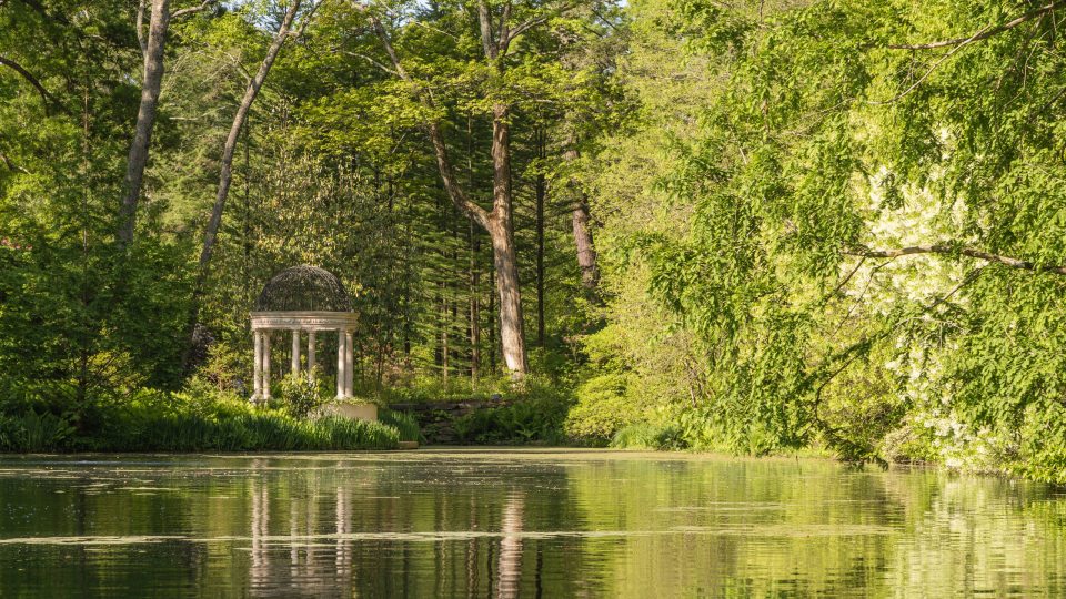 A small stone decorative structure is seen across a large lake surrounded by green trees