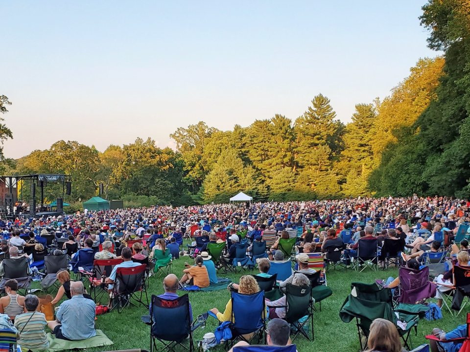 An outdoor concert overlooking a meadow with people sitting in chairs looking at the stage.