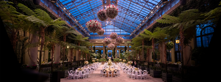 Long view of tables and chairs set up in an indoor conservatory setting for a formal event, beneath hanging baskets suspended from glass ceilings that are the deep blue color of the dusk beyond.