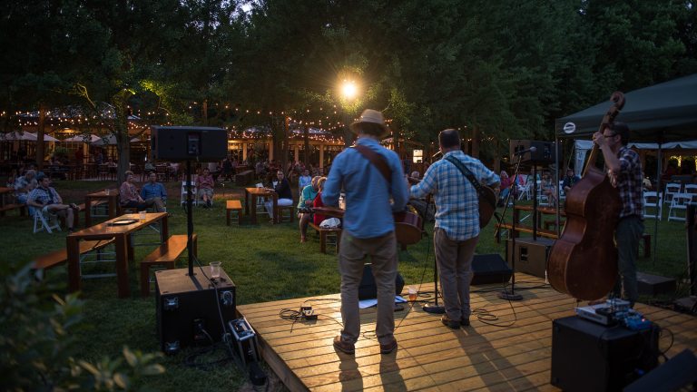 two men with their backs to the camera playing guitars on a wooden stage in an outdoor venue