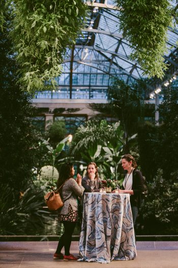 Three people with drinks stand in conversation around a tall round table in a lush Conservatory setting.