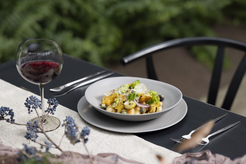 Closeup of a white bowl filled with food on a white plate on a black table set with silverware, a white runner, blue flowers, and a glass of red wine.