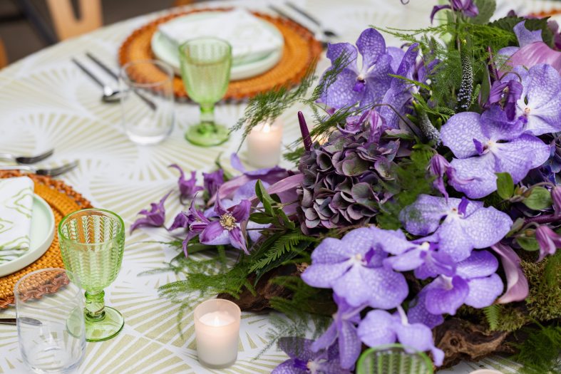 Closeup of table settings on a white and gold patterned tablecloth, with woven basket-like servers, round white plates, cloth napkins, green glass goblets, votive candles, and a floral arrangement in green and purple hues. 