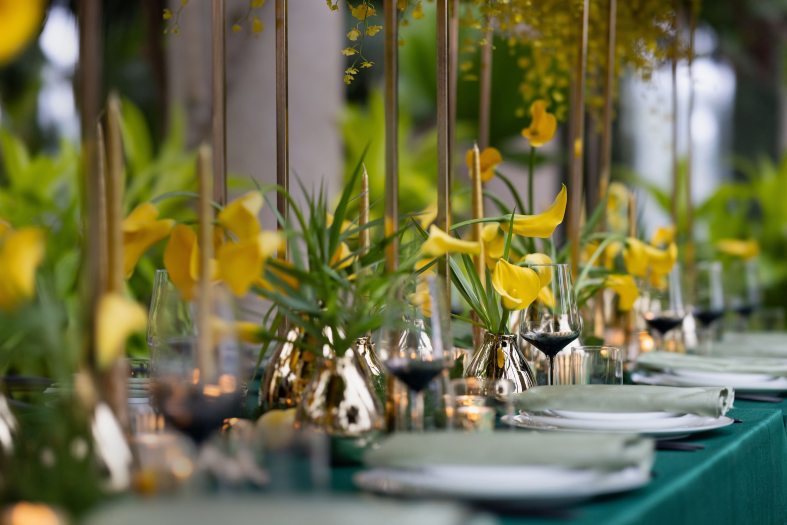 Diagonal shot of long table with dark green tablecloth and individual place settings decorated by silver vases of yellow calla lilies. 