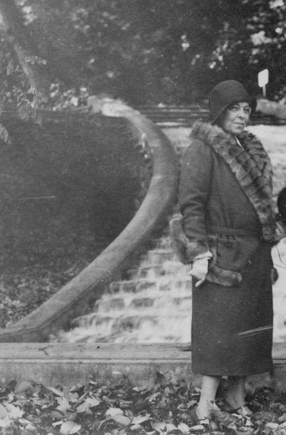 black and white image of a person in a long coat and hat looking at the camera