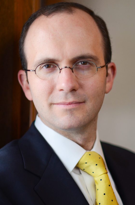 headshot of person with short dark hair in dark blue suit jacket, white shirt, and yellow tie; wearing oval wire-rimmed eyeglasses