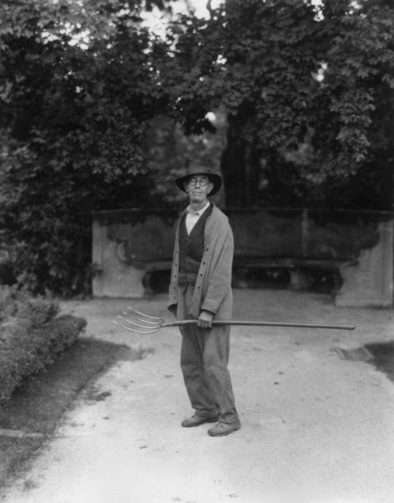A black and white image of a man holding a rake standing along a dirt path.