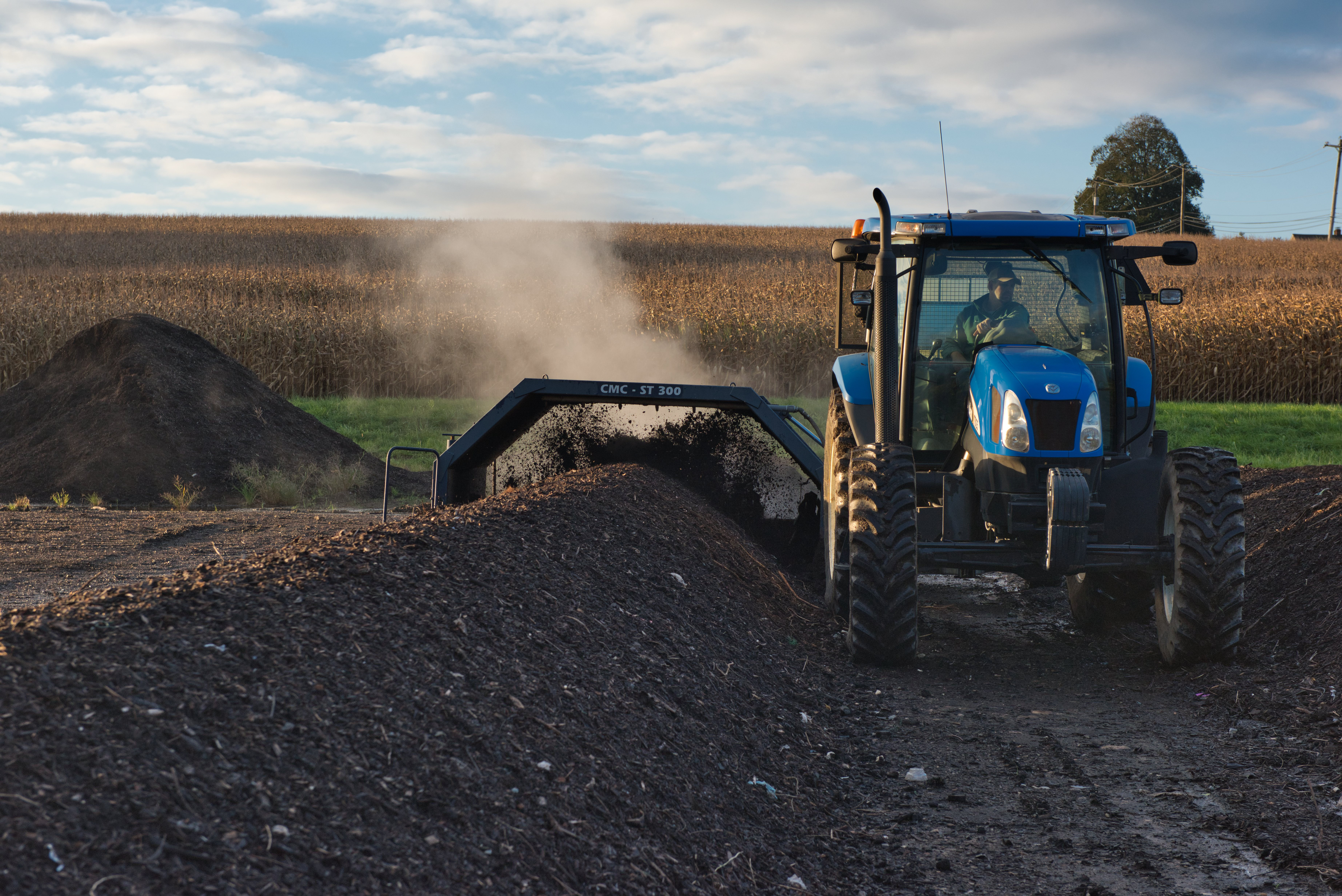 A large blue tractor is to the right of the image with a compost row in the foreground and fields with blue sky in the background. 