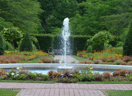 a round fountain set in the middle of two paths of gardens in bloom on either side of it