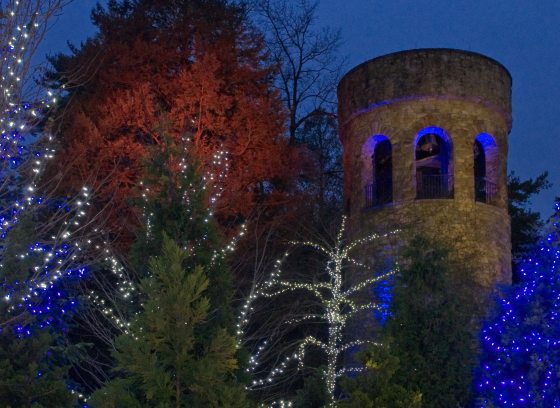 a stone tower with arched openings outlined in blue lighting against a darkening sky, with holiday-lit trees in the foreground