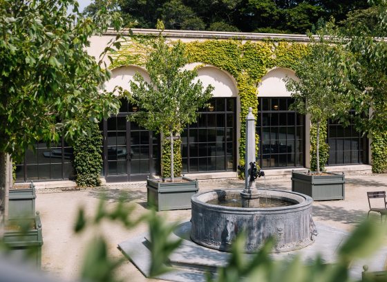 a circular fountain adorned with ornate sculptures sits in a courtyard surrounded by arching walls and green plants