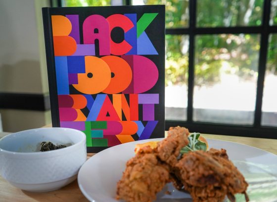Display of a white bowl filled with dark greens, and a white plate filled with fried food, in front of a book entitled "Black Food"