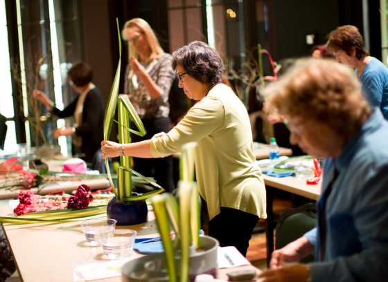 Several people standing at a table arranging floral greenery into vases.