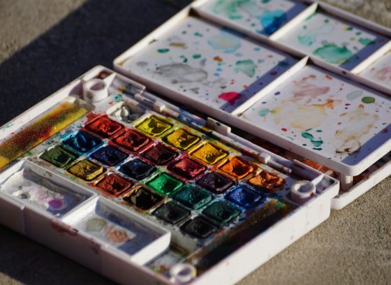A used box of watercolor paints, opened and laying on a tan surface.