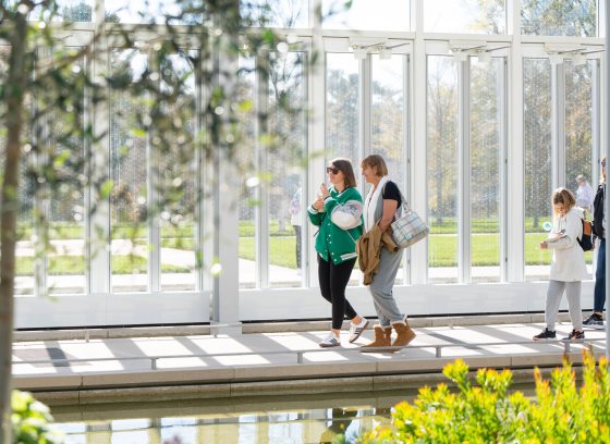 Guests stroll along a sunlit indoor path in a glass house, with water and green plants in view.