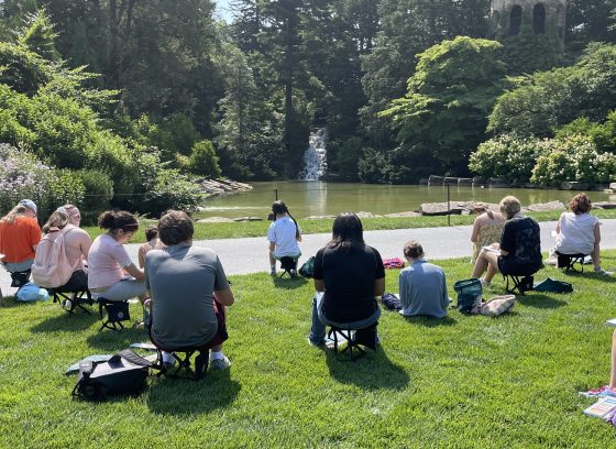 Young people, seated on folding garden chairs, sketch a landscape containing a pond, waterfall, and stone tower amid trees and gardens.