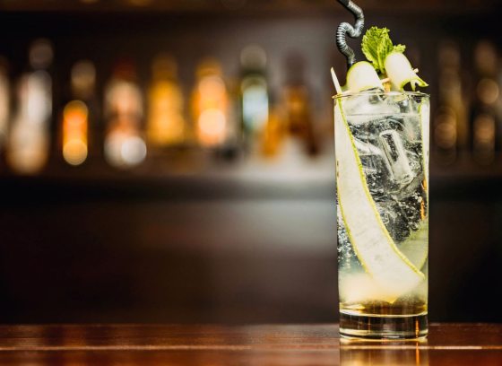 A clear cocktail, garnished with cucumber, onto of a dark wood bar. 