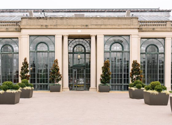 Wide view of conservatory exterior, with eight tall arched windows flanked by stone columns, and a series of large planters creating a broad pathway to the entrance.
