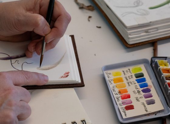 A person painting in a sketchbook with an open color palette next to them.