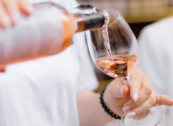 A close-up of a person pouring rosé wine into a clear wine glass at an outdoor event. The wine has a pink hue, and the person holding the glass has a light-colored shirt and manicured nails. The background is softly blurred, emphasizing the action of pouring.