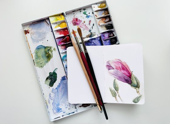 Watercolor paint palette with brushes and notebook containing a purple painted flower.