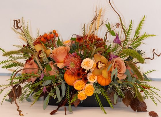 A fall themed centerpiece with flowers in ranges of yellow and orange set against a white backdrop.