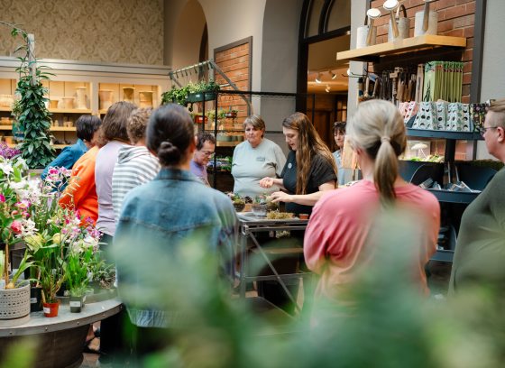 People in the Longwood Garden Shop, surrounding a table, watching someone pot a plant.