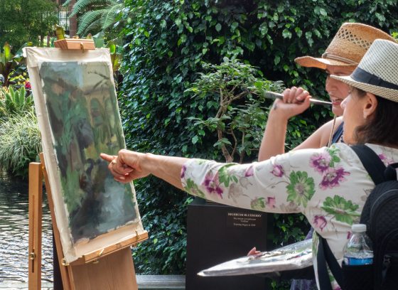 Two people looking and pointing at a painting in progress on an easel in a vegetated area.