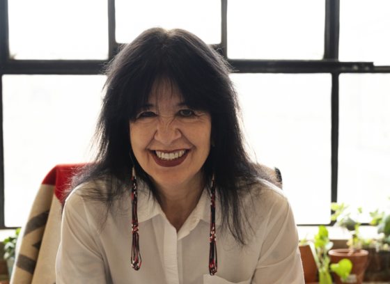 A person in a white shirt, black hair, with long beaded earring smiling at the camera.