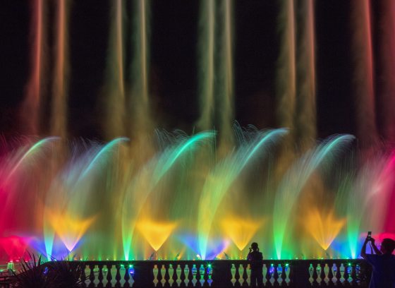 A row of parallel fountain pairs in rainbow colors shoot toward the sky, with a middle layer of dancing fountain spray, and a lower level of cone-shaped fountains.