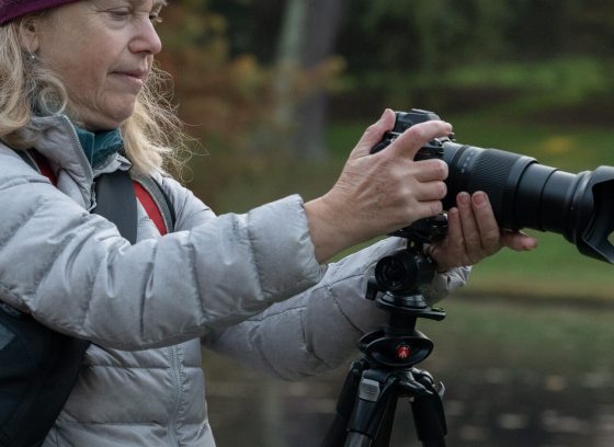 A person in a long coat and hat, taking a picture with a camera on a tripod.