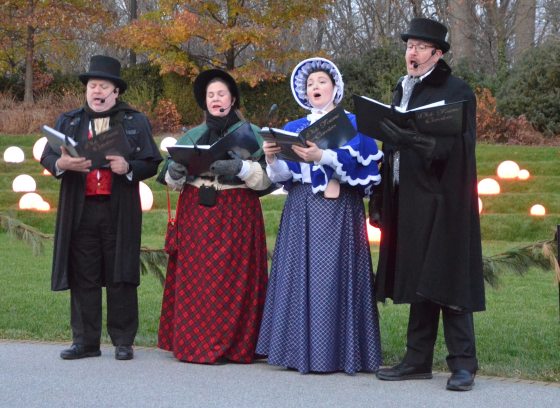 Four singers dressed in old-fashioned Dickens-era clothing and holding black songbooks, stand and sing in front of a lawn decorated with globes of white light.