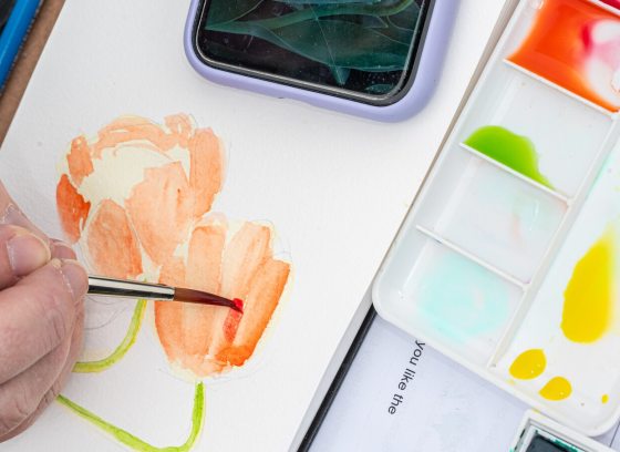 Close-up view of an artist painting an orange watercolor flower on paper, with a palette of vibrant paints and a smartphone visible nearby.