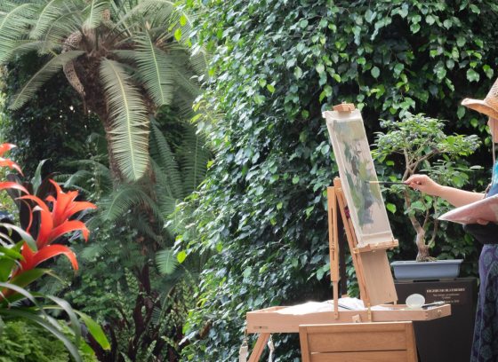 An artist wearing a straw hat paints a bonsai tree on an easel while surrounded by lush tropical plants and red bromeliads in a conservatory.