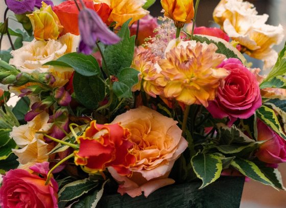 A close-up of a colorful, multi-layered bouquet with roses, tulips, and various blooms in shades of pink, yellow, orange, and purple.