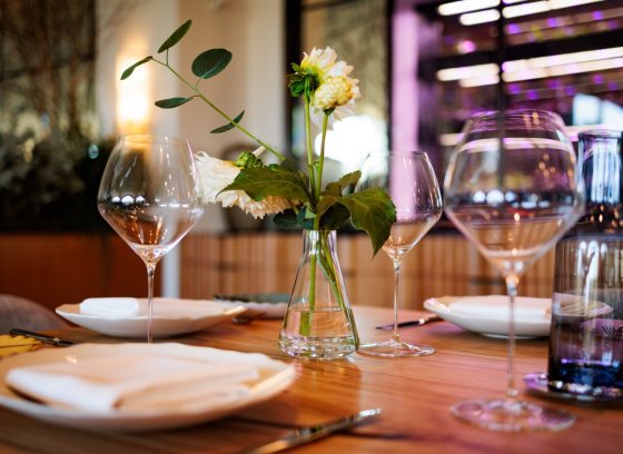 Set dining table with wine glasses, white plates, a napkin, a water bottle, and a small vase with a single white flower, suggesting a fine dining atmosphere.