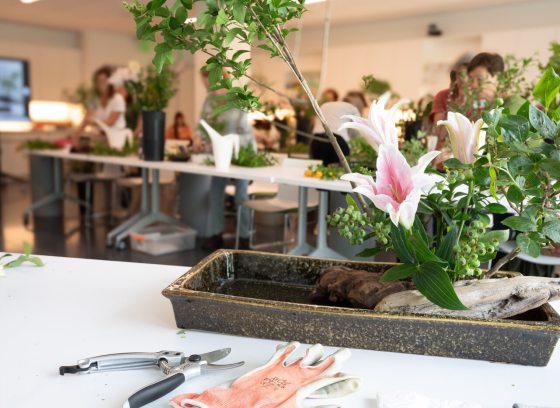An indoor floral design workshop with an arrangement in the foreground and other participants working at tables in the blurred background.