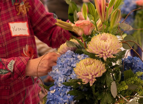 A person in a red plaid shirt creating a floral design with white and blue flowers.