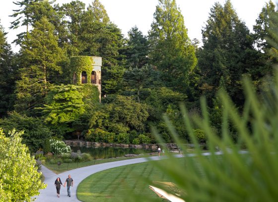 Two guests walk along a curved garden path amid lush greenery and a stone carillon tower in the background.