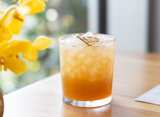 A peach drink, in a highball glass, set ona light wood table, with a vase of yellow flowers.