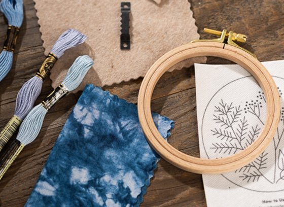 Embroidery supplies on a wood table, featuring a small hoop, thread in shades of blue, and a botanical illustration.