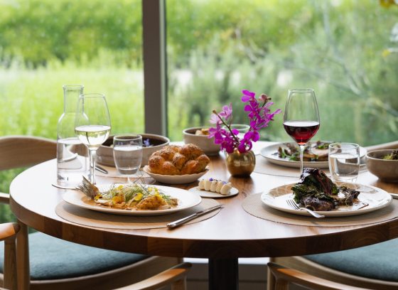 A table in a restuarant with plates of food and beverages, untouched, overlooking a green outdoor space.