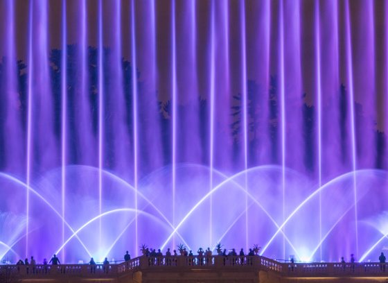 Fans and jets of fountain water bathed in lavender light rise above silhouettes of guests on a stone observation balcony.