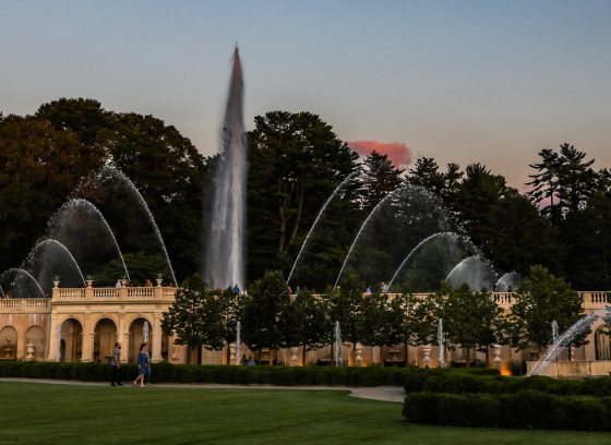 Wide view of a fountain garden at dusk, against the silhouette of trees and a clear deep blue sky.