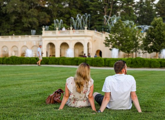 Two people relax on a green lawn while fountains dance in the background.