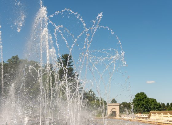 Fountain jets of dancing water rise up from a stone basin against a cloudless blue sky.