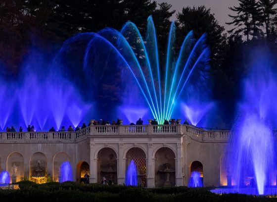 Fountains of water rise in hues of blue from the front and top of a stone facade.