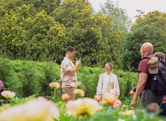 A guest with a smartphone captures the broad smile of another guest amid the blooms of a peony garden.